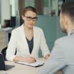 Woman in glasses interviews man at office desk.