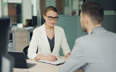 Woman in glasses interviews man at office desk.