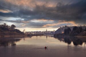Kayakiste pagayant seul sur une rivière calme avec une ville et ses églises en arrière-plan au coucher du soleil